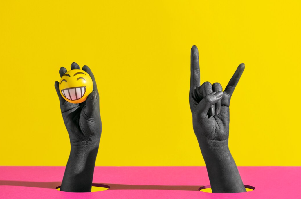 two black color hands, one holds  a ball with a smiling emoji on it on pink and yellow background