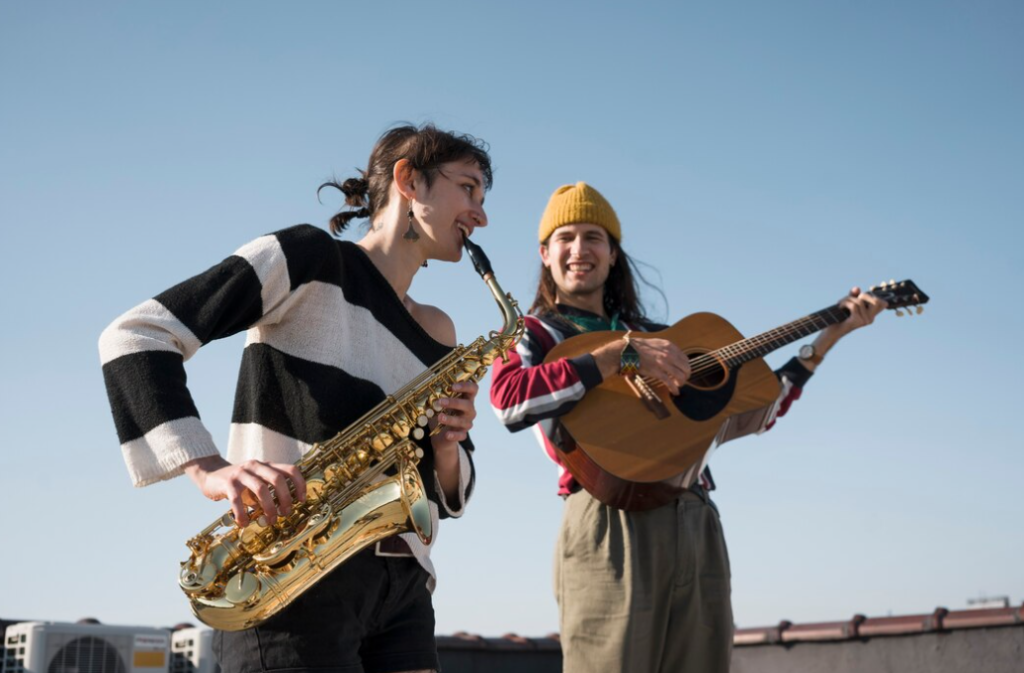 a woman plays a saxophone, while a man with a beanie strums a guitar against a clear sky