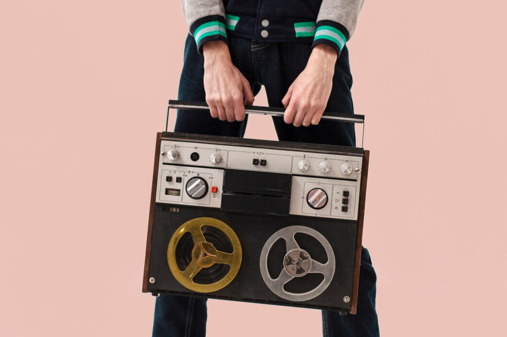 A person holds a vintage reel-to-reel tape recorder against a pink background