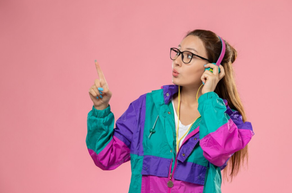 woman in colorful jacket pointing up while listening to headphones on a pink background