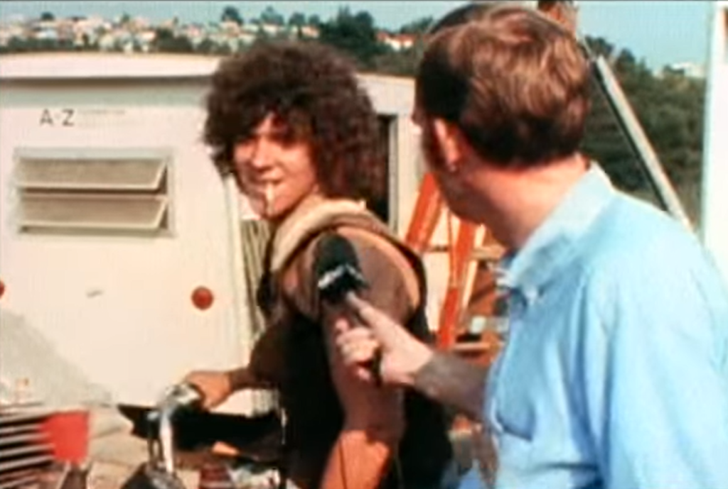 Two individuals, one with curly hair, stand by a white vehicle with the label "A 2"